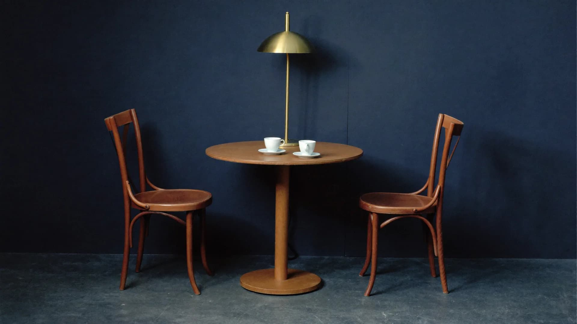 Two empty chairs at a small round table under one brass lamp, two cups waiting