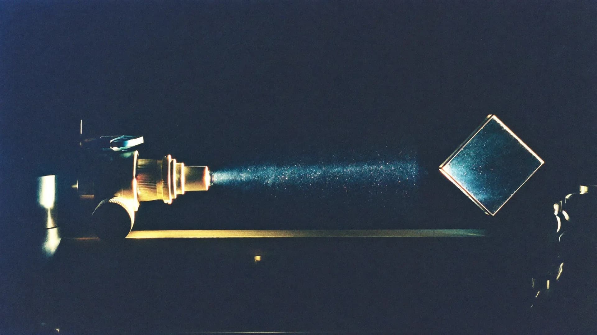 A brass projector lens on an optical bench casting one dusty blue beam onto a tilted square plate