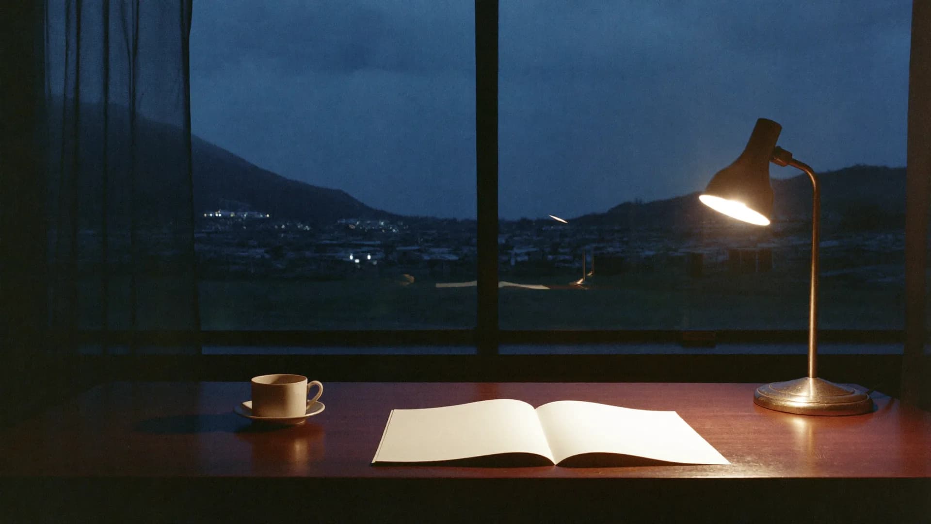 A desk by a wide window at dusk: one lamp lit, an open blank notebook and a cup, town lights far below