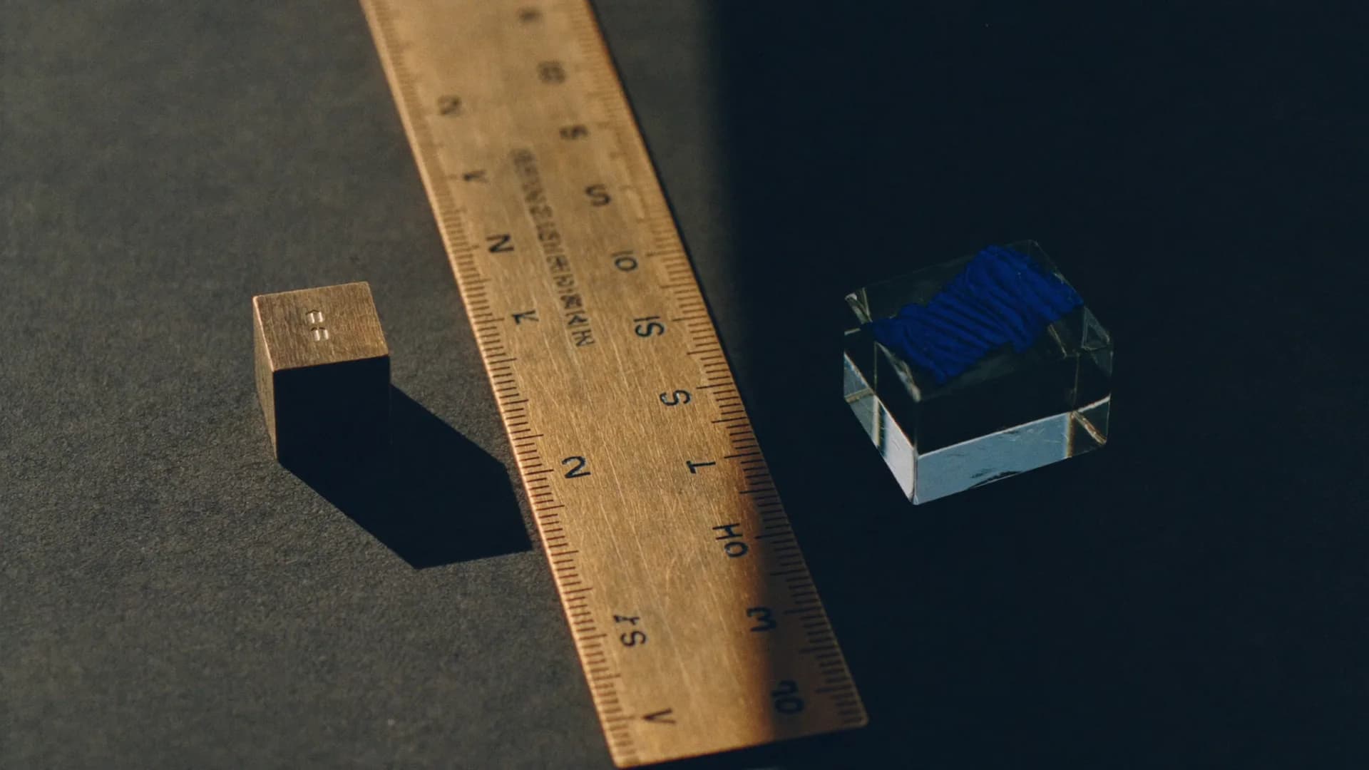 A brass ruler between a small brass cube and a glass cube, a hard edge of light splitting the frame